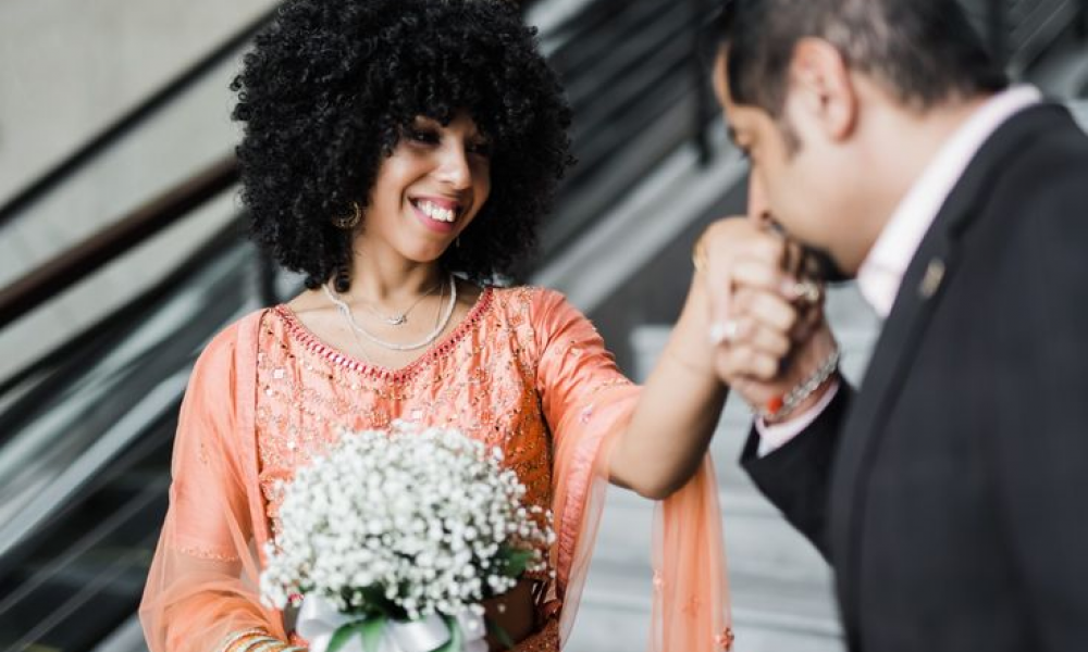 A joyful couple stands on stairs celebrating their engagement or wedding moment together. The woman wears an elegant peach dress with beautiful embroidery while holding a bouquet of white flowers. The man in dark suit kisses her hand displaying affection against a festive backdrop creating a memorable scene filled with love.