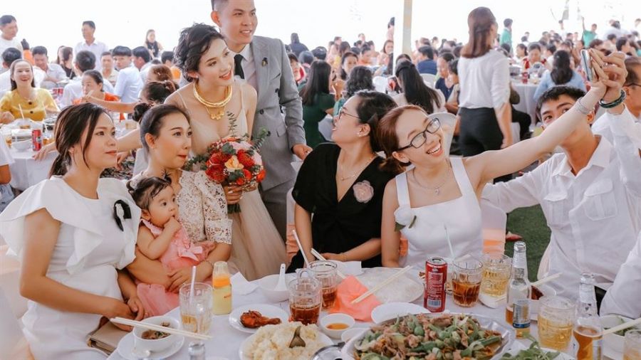 A lively wedding reception scene in Dubai featuring a joyful bride in an elegant gown and a groom stylishly dressed. Guests are seated at tables enjoying themselves, some taking selfies. A table full of gourmet food is visible, enhancing the festive atmosphere. Bright lights fill the venue showcasing a happy daytime celebration filled with love and cheer.