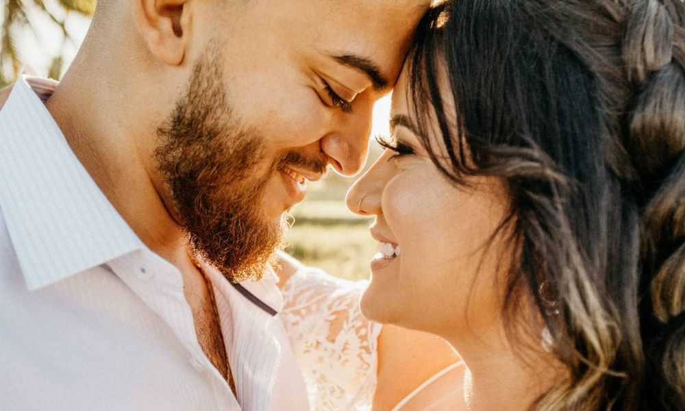 A happy couple leans together with their foreheads touching in an outdoor setting filled with soft natural light. The man has a beard and wears a light shirt while the woman in white lace shows her braided hair cascading down her back. Their smiles radiate warmth and love against a softly blurred background, encapsulating the joy of sharing their special moment together on their wedding day.
