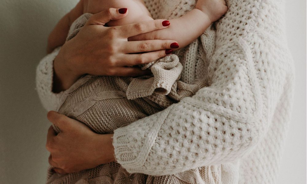 The image depicts a close-up of an adult holding a baby tightly in a cozy embrace. Both are dressed in soft knit garments in neutral colors with the adult displaying well-groomed red nails. The baby is swaddled in a knitted blanket, showcasing warmth and affection. The minimalistic background highlights this loving connection beautifully, creating a soothing atmosphere that emphasizes their bond.