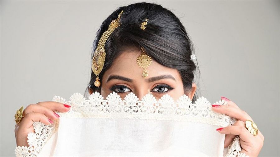 A woman partially covers her face with a delicate white lace fabric while revealing expressive eyes highlighted by dark eyeliner. Her beautifully styled hair is adorned with traditional gold jewelry including a maang tikka on her forehead. She holds the fabric with red manicured nails that add color to her soft look against a neutral background, showcasing elegance and cultural elements present in wedding traditions.