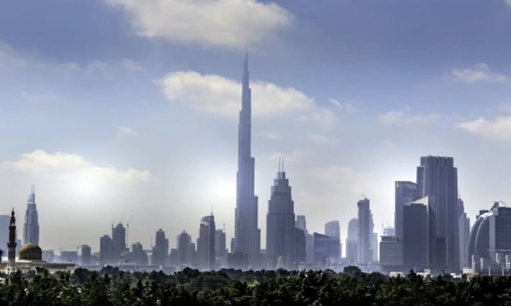 The image shows a beautiful view of Dubai's skyline featuring the Burj Khalifa surrounded by modern skyscrapers. The foreground has lush greenery which contrasts with the urban structures in the background. The sky is partly cloudy creating soft shadows on the buildings while highlighting their futuristic designs. This panorama captures Dubai’s dynamic essence as a global hub blending nature with impressive architecture offering a glimpse into its vibrant lifestyle.