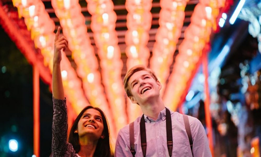 A happy couple stands together under bright red lanterns glowing softly around them. The woman joyfully points upward while the man smiles broadly at her side. They appear relaxed and casual as they share a delightful moment together in a festive atmosphere. The surrounding lights create a warm ambiance that highlights their happiness and love for each other amidst the vibrant scene.