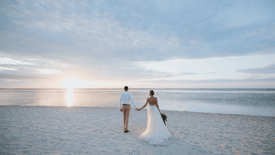A couple walks hand in hand on a serene beach during sunset. The bride’s flowing white gown contrasts beautifully with the groom's light-colored outfit. Soft clouds float across the sky as they approach the warm ocean water, creating an ambiance of romance and tranquility perfect for weddings or intimate moments. The wide sandy beach surrounds them while the colors of sunset illuminate this peaceful scene inviting couples to create their own wedding dream here in Dubai.