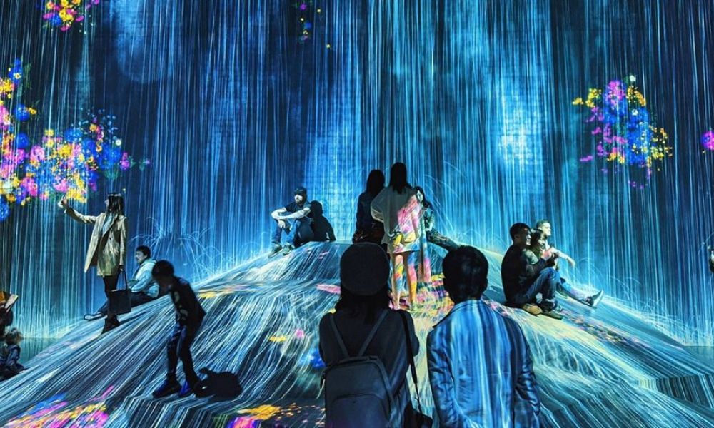 The image shows people on a vibrant digital hill surrounded by colorful lights and patterns. They are enjoying an interactive experience that combines technology and creativity in a futuristic setting.