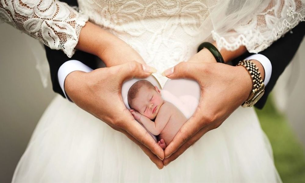 A couple dressed in wedding attire forms a heart shape with their hands, symbolizing love and unity. The woman wears a beautiful lace dress, while the man is in a sharp dark suit. The background features soft greenery that adds romance to the scene. Inside their heart-shaped hands is an edited image of a sleeping baby, representing family and new beginnings.