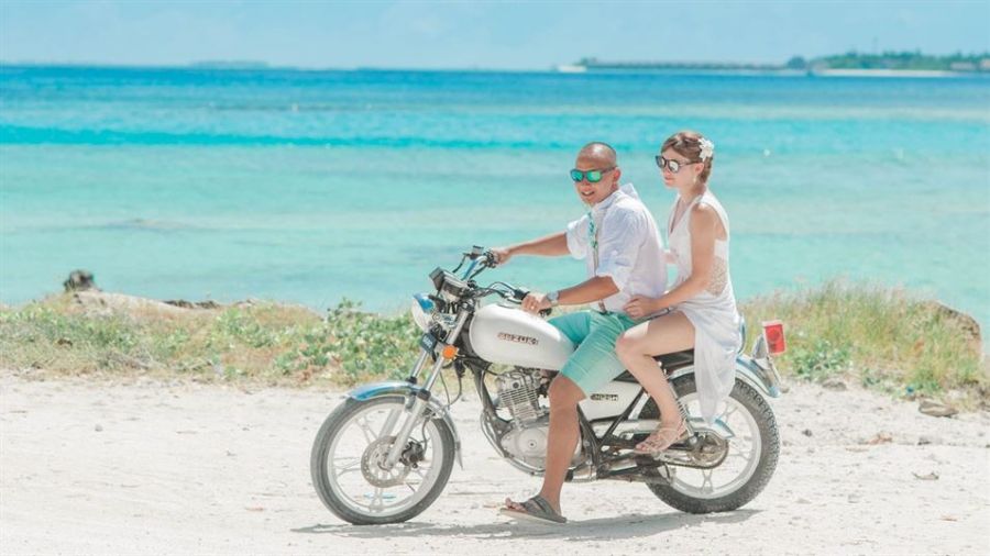 A happy couple rides a motorcycle along a beautiful beachfront in Dubai. They wear sunglasses and summery clothes, with turquoise ocean waters in the background under a bright blue sky. The sandy path beside them has scattered rocks and sparse plants, creating a peaceful atmosphere that speaks of adventure and relaxation.
