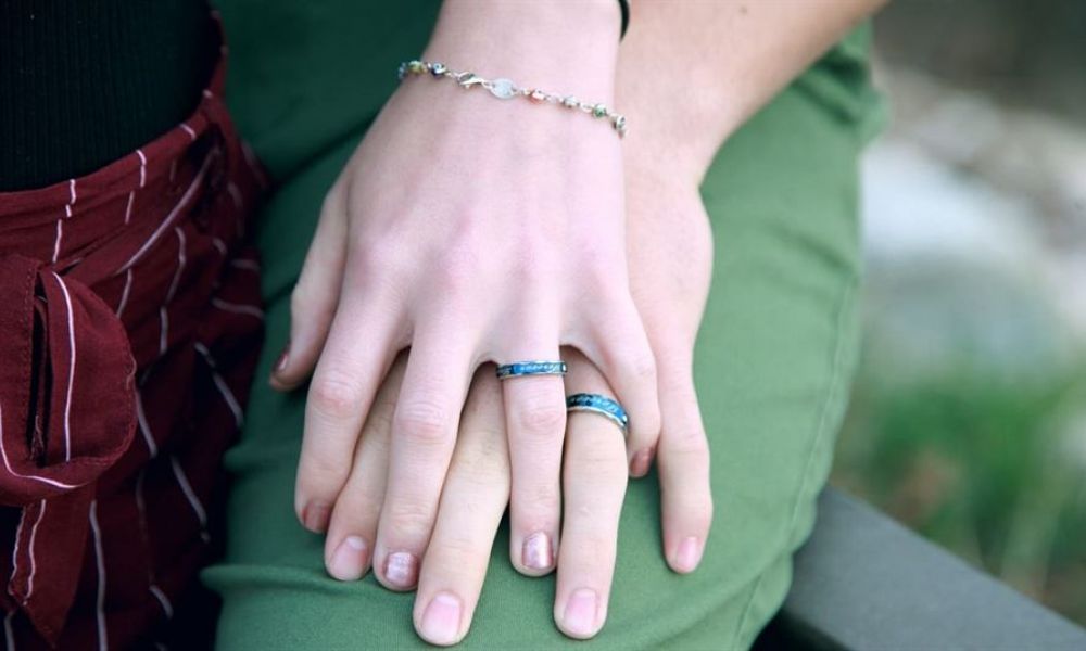 The image features two hands gently resting on top of each other. One hand wears a delicate silver bracelet and a blue stone ring, while the other also has a similar blue stone ring. The background is softly blurred, enhancing the focus on their connection, which symbolizes unity and companionship in love. A maroon, pinstriped fabric is visible near one wrist, indicating casual attire while adding warmth to the scene.
