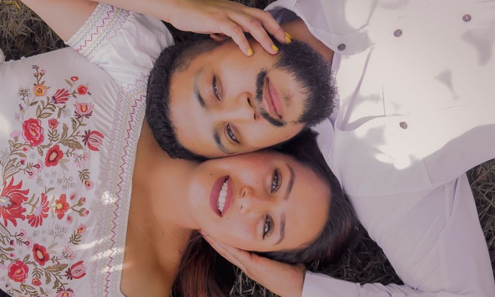 The image shows a couple lying together on the grass, enjoying a serene moment under gentle sunlight. The man in a white shirt has a beard while the woman in an embroidered dress shows off her long hair and bright smile. They look happy and relaxed, creating an intimate setting that feels peaceful and romantic.