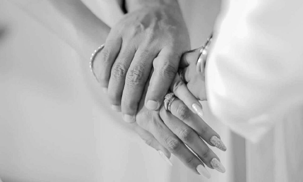 The image features two hands gently holding each other, symbolizing love and commitment. One hand has beautifully manicured nails adorned with a sparkling ring, while the other seems larger and protective. The couple wears light-colored clothing, creating an elegant atmosphere. This intimate moment illustrates the beauty of union during a wedding ceremony.