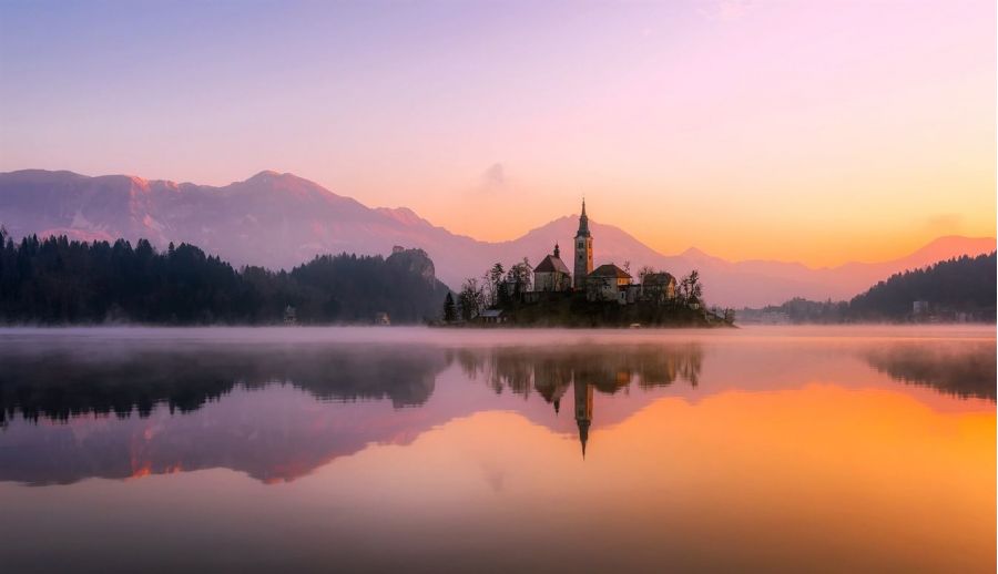 The image shows a beautiful island at sunrise featuring a picturesque church surrounded by calm waters. Mist rises from the water creating an enchanting atmosphere. Silhouetted trees frame the scene while majestic mountains glow softly in pink and orange hues beyond them. The landscape conveys peace and natural beauty perfect for weddings or romantic escapes.