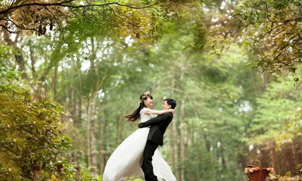 An excited bride and groom are celebrating in a lush forest setting surrounded by vibrant greenery. The groom is wearing a black suit while the bride shines in a flowing white dress decorated with flowers. They radiate happiness as they embrace each other joyfully, with a picnic basket nearby adding charm.