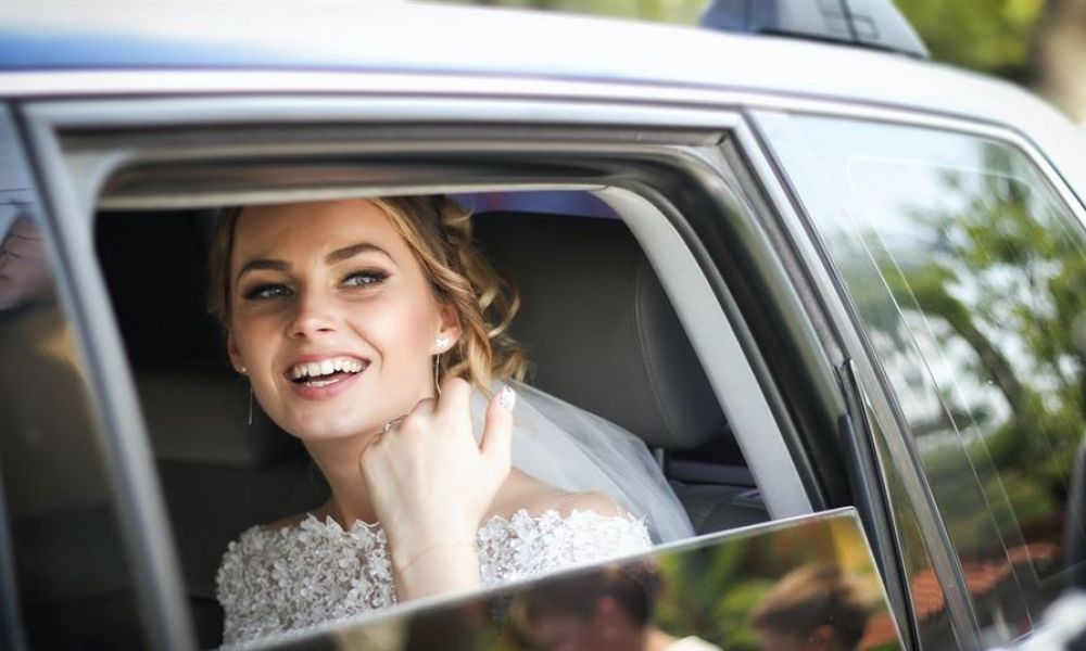 7932F836 | Registering For The Seychelles Wedding In Dubai! | You Will Need All The Documents That We Have Listed Above And You’ll Need To Bring Them To The Mofa (Ministry Of Foreign Affairs). The Ministry Of Foreign Affairs Will Stamp Your Seychelles Wedding Certificate That Will Then Be Recognizable By The Government. A joyful bride sits in a spacious car, wearing an elegant lace-embroidered wedding dress. She has a radiant smile and her hair is styled with a veil flowing down her back. The warm sunlight filters through the window, creating a happy atmosphere. The background features blurred greenery, adding a romantic touch to her special moment.