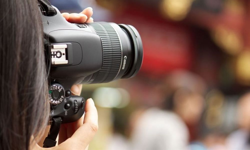 A person is holding a DSLR camera focusing on capturing a scene. The camera's details are visible while the background shows colorful blurs creating a lively atmosphere that represents an outdoor setting.