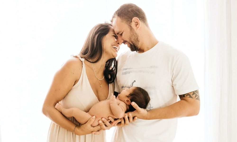 A happy couple stands close in a soft-lit room holding their newborn baby joyfully. The woman wears a light peach dress while the man sports a white shirt with tattoos visible on his arms, radiating warmth and love.