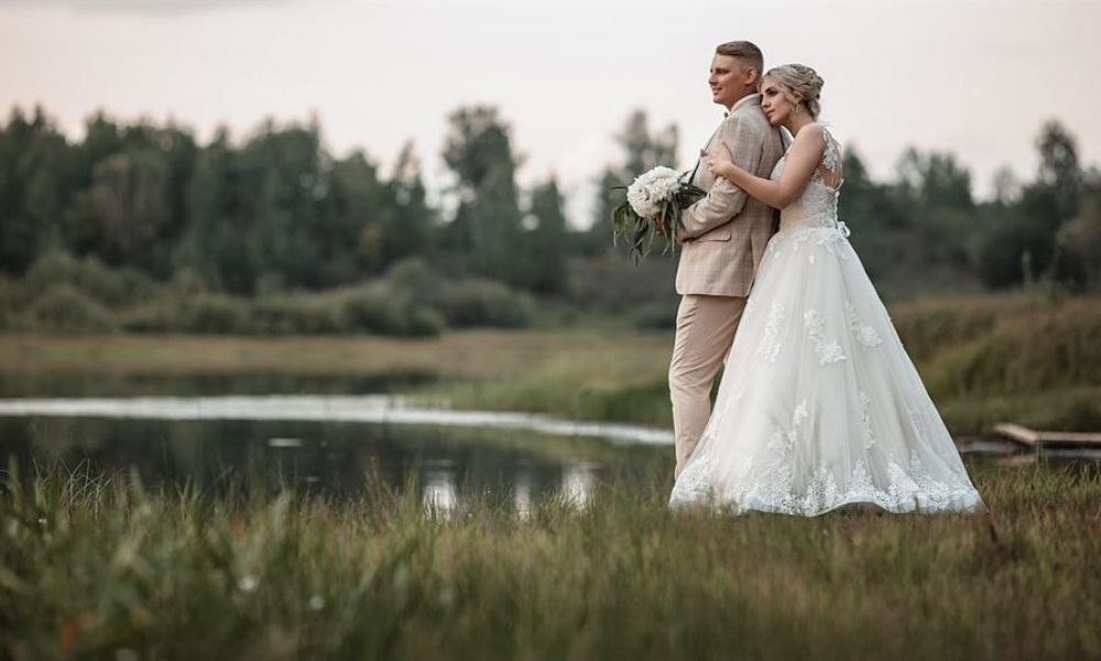 A lovely couple stands by a serene lake, surrounded by lush greenery. The bride wears an elegant white gown adorned with lace, while the groom looks dapper in a beige suit. They stand peacefully, with the bride gently embracing the groom from behind, holding a bouquet of white flowers. The background features soft-focus trees and a cloudy sky which adds to the romantic feeling of this tranquil scene. This captures their tender moment on their special day amidst nature's beauty.