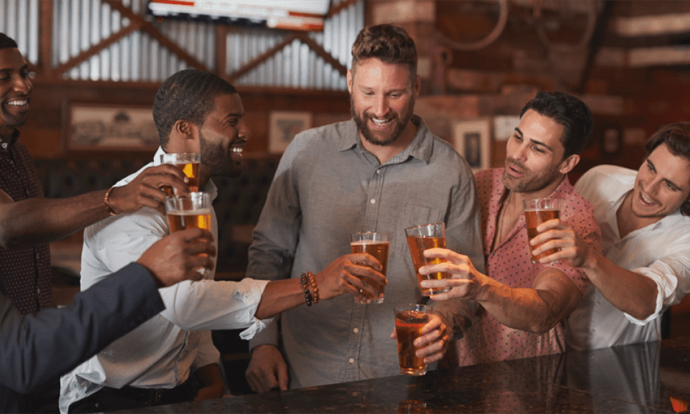 A fun group of five men at a bar are joyfully raising glasses filled with beer for a toast. They are dressed casually and appear relaxed amidst a lively atmosphere decorated with wooden panels and cheerful pictures, suggesting a celebration like a bachelor party.