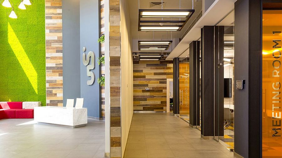 A bright office interior with a vertical garden wall, minimalist white furniture, gray tiled floor, glass-walled meeting rooms labeled Meeting Room 1, wood paneling, orange accents, and sleek lighting fixtures creating an uplifting atmosphere.