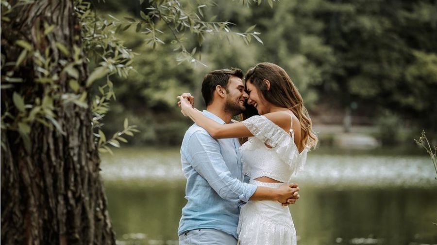A loving couple embraces by a serene lake, creating a romantic scene surrounded by lush greenery. The man wears a light blue shirt while the woman looks beautiful in her white lace dress, symbolizing love and commitment. This idyllic outdoor setting enhances their affectionate moment together, portraying warmth and tranquility that resonates with every couple's special day.