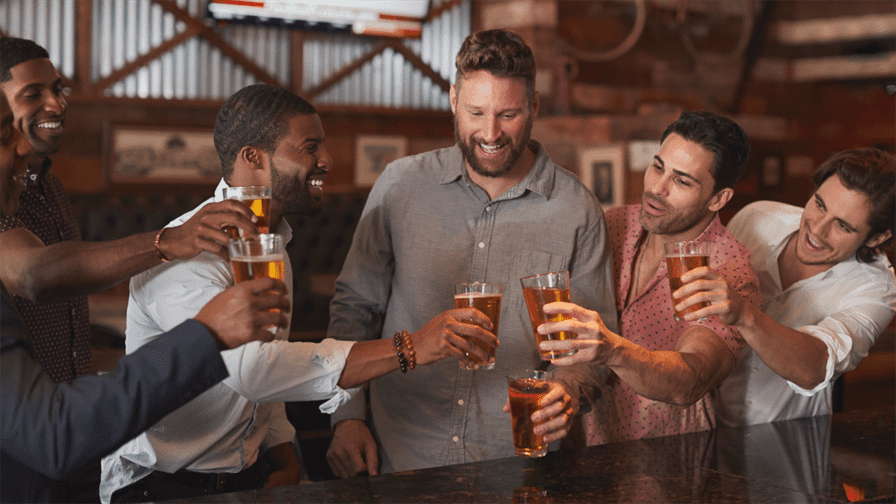 A fun group of five men at a bar are joyfully raising glasses filled with beer for a toast. They are dressed casually and appear relaxed amidst a lively atmosphere decorated with wooden panels and cheerful pictures, suggesting a celebration like a bachelor party.