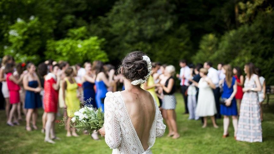 The image shows a bride in a lace dress holding a bouquet of white flowers facing away from the camera. Guests are mingling on lush green grass, enjoying the festivities of an outdoor wedding. The ambiance is vibrant with colorful attire worn by attendees who celebrate joyfully in a beautiful garden setting, embodying romance and happiness during this special occasion.