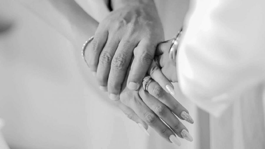 The image features two hands gently holding each other, symbolizing love and commitment. One hand has beautifully manicured nails adorned with a sparkling ring, while the other seems larger and protective. The couple wears light-colored clothing, creating an elegant atmosphere. This intimate moment illustrates the beauty of union during a wedding ceremony.