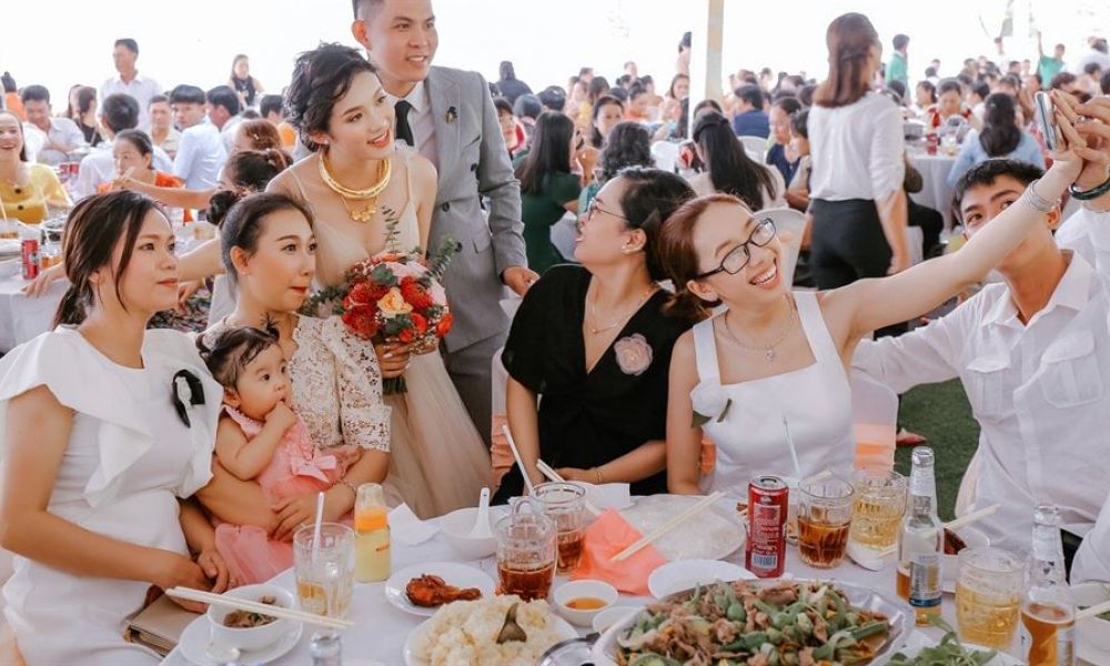 A lively wedding reception scene in Dubai featuring a joyful bride in an elegant gown and a groom stylishly dressed. Guests are seated at tables enjoying themselves, some taking selfies. A table full of gourmet food is visible, enhancing the festive atmosphere. Bright lights fill the venue showcasing a happy daytime celebration filled with love and cheer.