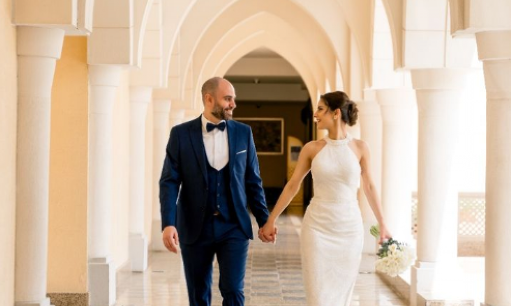 A1D343Ba | Government Launches 75,000 Aed Abu Dhabi Rental Support Initiative: All You Need To Know | The Uae Government Has Just Announced A Groundbreaking Initiative That Is Sure To Aid Its Residents. An elegant couple is happily walking hand in hand down a grand corridor. The woman shines in her white gown while the man looks dapper in his blue suit. Intricate architecture surrounds them. Soft light enhances their joyful expressions as they share a loving glance amidst the sophisticated decor. This scene beautifully captures the essence of romance and celebration perfect for any wedding day.