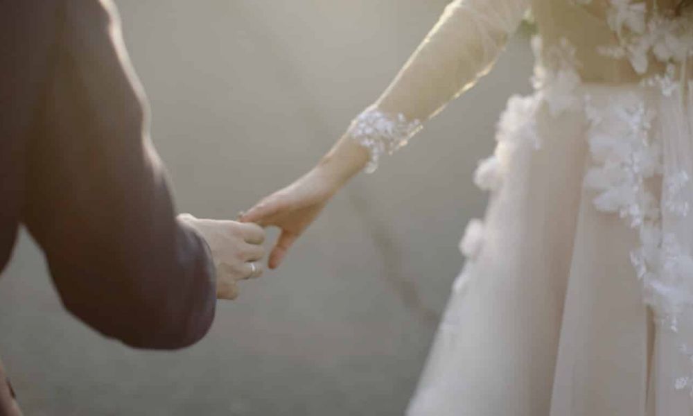 Easy wedding guide for Oman expats a510847b | Easy wedding guide for Oman expats in UAE | The wedding day is a dream for every couple. But getting married in any GCC country including Dubai can be a hectic and long process. Dubai is home to some extraordinary luxurious wedding venues but these can cost a fortune and can be near impossible to secure. There are several options to have a legal wedding in Dubai as long as the marriage is registered in the court or the relevant embassies. A close-up image of a couple holding hands, with one partner in a dark suit and the other wearing an elegant white wedding dress decorated with floral lace details. The setting is softly lit with warm sunlight, giving a romantic feel, while the background is blurred to focus on their connection. This image symbolizes love and partnership during wedding ceremonies in Dubai or Abu Dhabi.