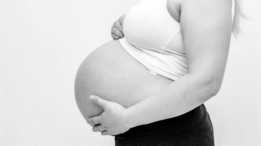 A black-and-white photograph shows a pregnant woman’s side profile wearing a fitted white tank top and dark skirt. Her right arm cradles her baby bump while her left arm supports the bottom of her belly. The light-colored background emphasizes her form, capturing a serene moment that highlights themes of motherhood and anticipation without showing her face or surroundings.