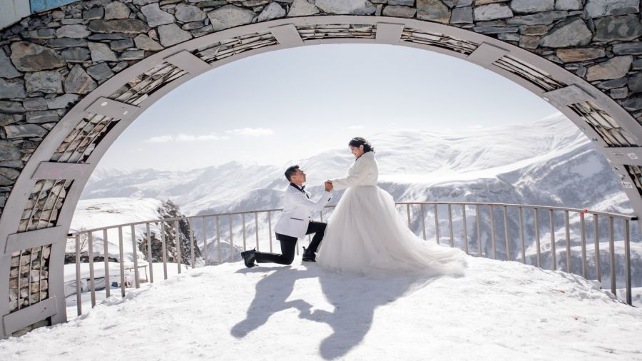 A romantic image featuring a couple in snowy mountains. The bride wears a flowing white dress with a matching jacket while the groom is on one knee holding her hand under an arched stone structure. Snow-covered peaks and bright skies create an enchanting backdrop, capturing a magical moment of love and commitment.