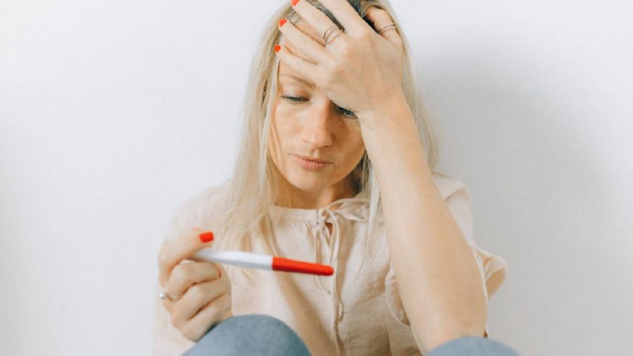 A woman sitting against a white background looks concerned while holding a pregnancy test in one hand. She has blonde hair, wears a light-colored blouse, and has red nail polish on both hands. The image captures her deep thought as she reflects upon personal circumstances surrounding the test result during an emotional moment.