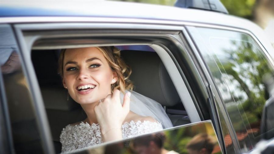 A joyful bride sits in a spacious car, wearing an elegant lace-embroidered wedding dress. She has a radiant smile and her hair is styled with a veil flowing down her back. The warm sunlight filters through the window, creating a happy atmosphere. The background features blurred greenery, adding a romantic touch to her special moment.