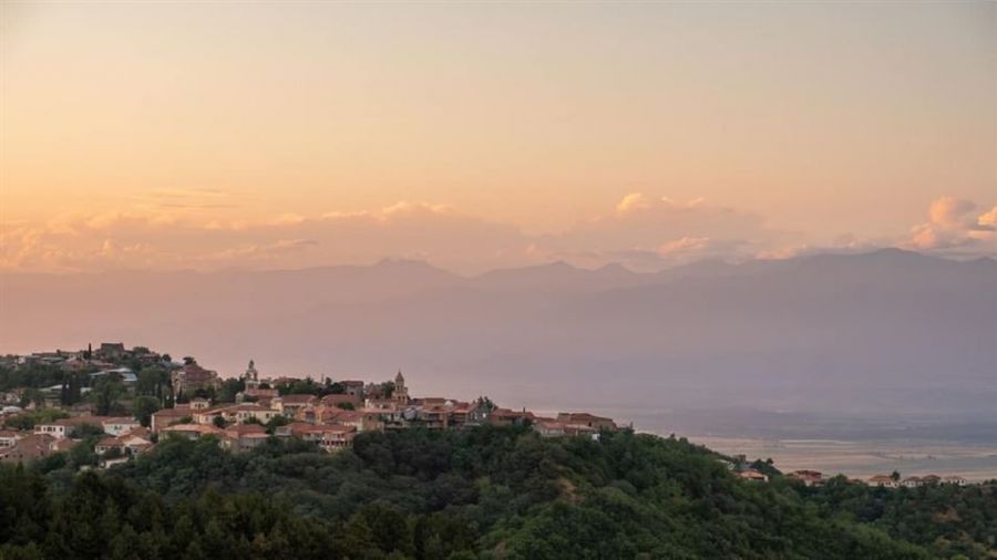 A beautiful sunset over a quaint town nestled in lush green hills showcases charming earth-toned buildings and distant mountains under a soft orange-pink sky, creating a serene atmosphere ideal for weddings and ceremonies.