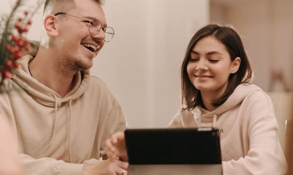 A young couple relaxes together while looking at a tablet screen in a cozy setting. The man wears glasses and smiles cheerfully while the woman sits beside him with shoulder-length dark hair, sharing smiles over the device. Warm lighting enhances their relaxed atmosphere, surrounded by softly blurred plants that add intimacy to this joyful moment.