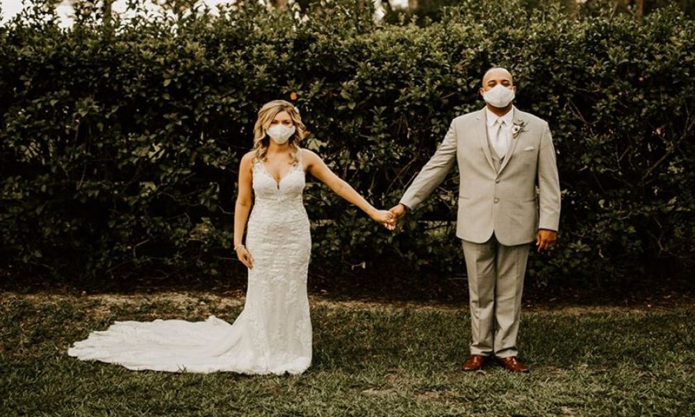 A happy couple stands outside holding hands surrounded by lush greenery. The bride is wearing a beautiful white lace gown while the groom is dressed in a light grey suit. They are both wearing masks, symbolizing love during challenging times. Warm lighting enhances the serene setting, perfect for an intimate wedding ceremony that looks magical and memorable.