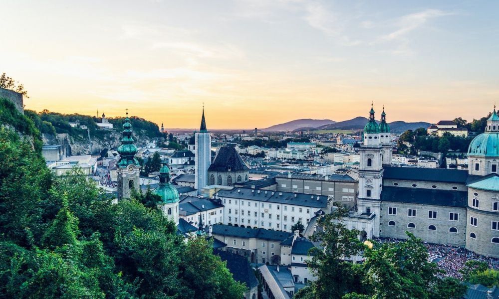 A beautiful sunset over a cityscape showcasing historic architecture with domed roofs and church steeples. On the left, a lush hillside is framed against the warm, colorful sky filled with orange and pink hues as dusk approaches. The urban scene reflects tranquility and timeless beauty at this unique wedding destination perfect for creating memorable ceremonies in Dubai or Abu Dhabi.
