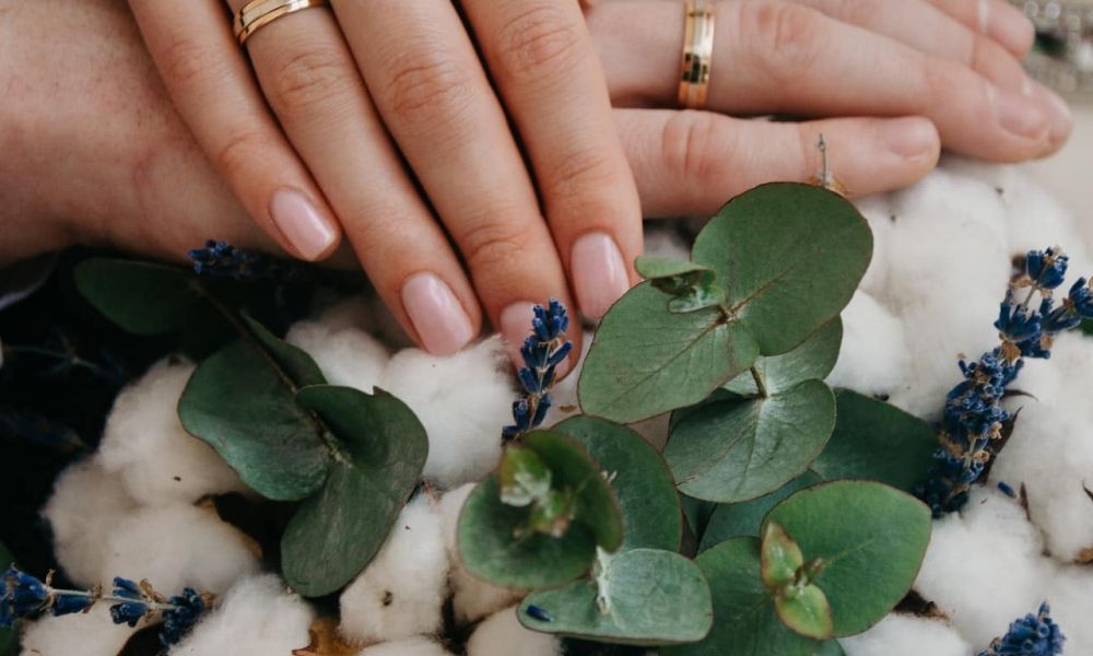 This image shows two hands gently resting on soft white flowers, both wearing beautiful wedding rings. The hands are surrounded by green eucalyptus leaves and delicate blue flowers, all bathed in warm light that creates an atmosphere of love and commitment. The composition symbolizes union and celebration against a natural backdrop, evoking feelings of intimacy and warmth.