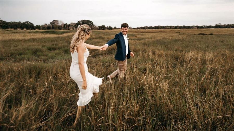 The image showcases a happy couple enjoying nature together in wide open grasslands under a cloudy sky. The woman wears a beautiful white dress while the man is dressed smartly in blue and light pants. They are holding hands, reflecting love and joy as they share this moment outdoors. The serene setting adds charm to their adventure-like appearance amidst tall grass and gentle vegetation.