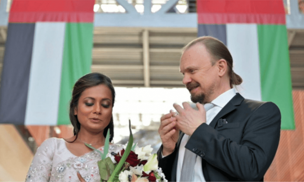 A happy couple stands under two United Arab Emirates flags inside a beautifully decorated venue. The woman holds a bouquet of flowers and wears an elegant dress while the man is in a smart suit. Both share a joyful moment as they look at something together, possibly an engagement ring, surrounded by celebration and love.