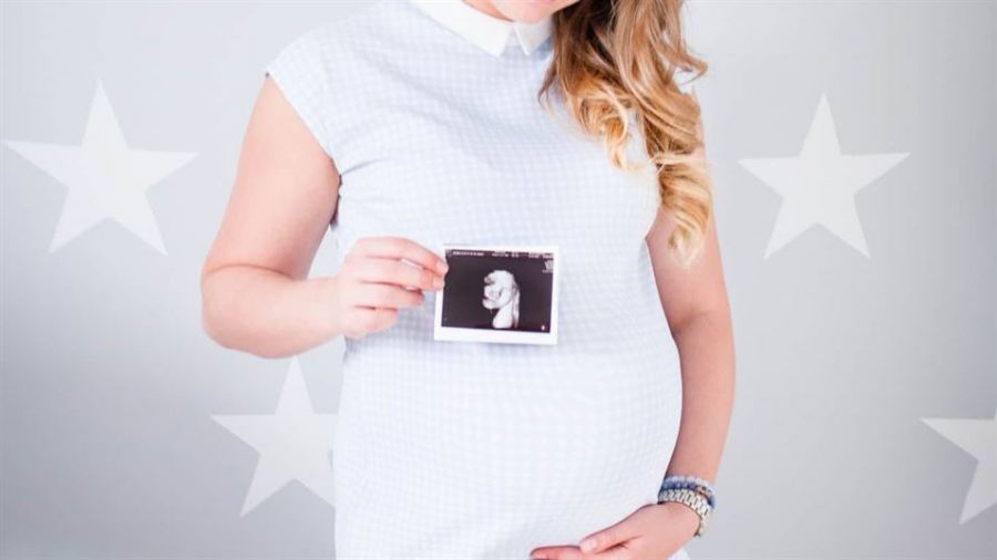 The image features a pregnant woman wearing a light blue dress holding an ultrasound photo in front of her belly. She has loose wavy hair, adorned with several bracelets on her wrist. The background displays large white stars over a gray backdrop, enhancing the theme of motherhood and anticipation for new beginnings. This picture captures emotions of excitement and family warmth as she dreams about the upcoming arrival of her child.