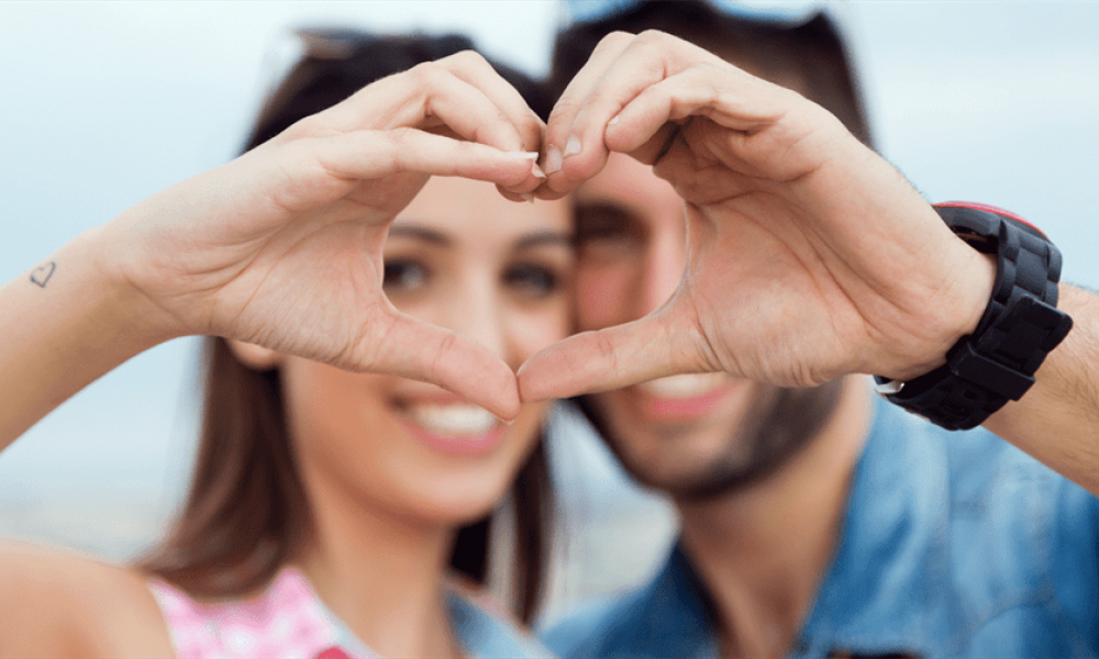 The image shows a happy couple forming a heart shape with their hands against a blurry background. They are smiling and casually dressed, radiating joy and love. The woman has long hair with sunglasses on her head while the man also has sunglasses pushed back. This romantic moment emphasizes the beauty of connection between them as they celebrate their love story in Dubai.