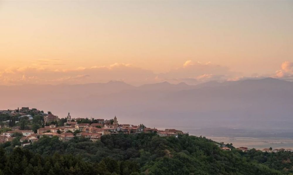 A beautiful sunset over a quaint town nestled in lush green hills showcases charming earth-toned buildings and distant mountains under a soft orange-pink sky, creating a serene atmosphere ideal for weddings and ceremonies.