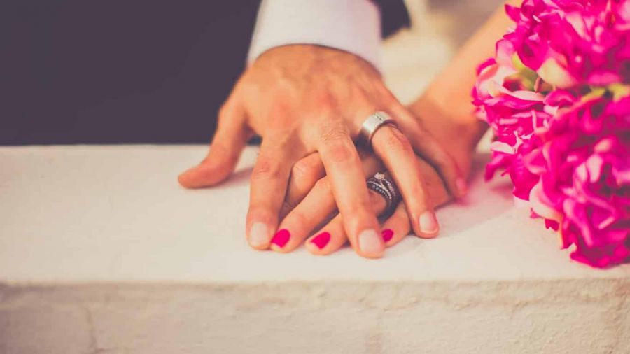A romantic scene featuring two clasped hands adorned with rings resting on a white surface. One hand has bright red polished nails while next to them lies a bouquet of vibrant pink flowers. The image captures themes of love and commitment in an intimate setting with warm lighting that emphasizes the joy of getting married. This visually speaks to couples considering their wedding options in Dubai and Abu Dhabi for their big day.