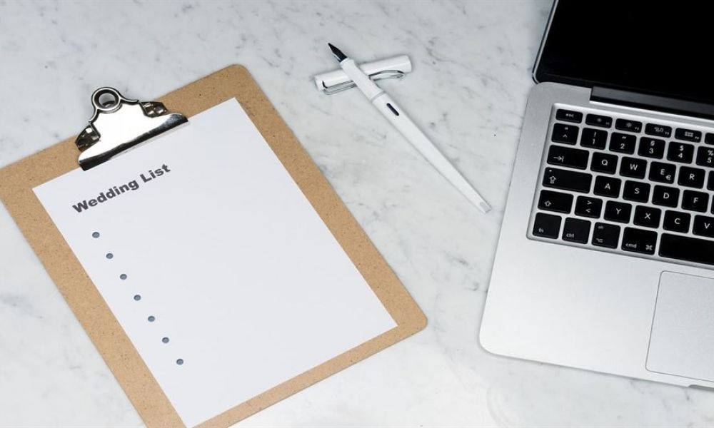 The image shows a clipboard labeled Wedding List on a marble surface next to an open laptop. A pen is placed between them, suggesting someone is planning their wedding preparations. The clipboard appears ready for jotting down ideas or tasks needed for organizing the wedding event effectively. The overall look inspires excitement and readiness for the big day ahead.