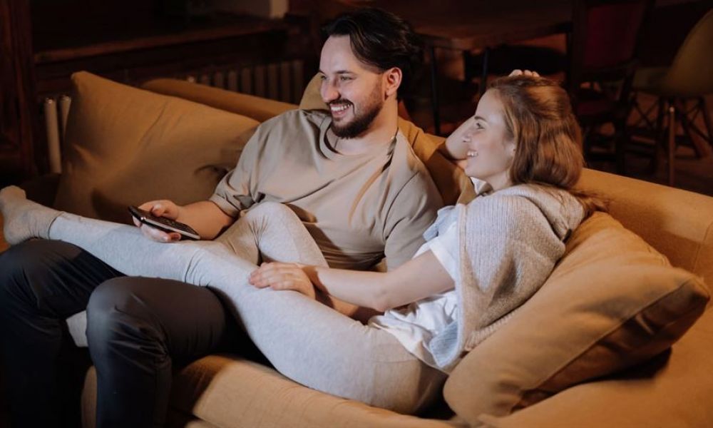 A happy couple is sitting on a light brown couch enjoying a cozy moment together. The man wears a light brown shirt while the woman is dressed in a comfy gray sweater. They appear relaxed and connected in their warmly lit living room, creating an inviting atmosphere perfect for couples considering marriage in Dubai.