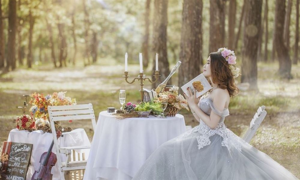 The image shows a bride wearing a light blue gown sitting at an elegantly set table in a peaceful forest. The table features flowers, candelabra, and champagne glass while soft sunlight illuminates the scene creating a serene atmosphere fitting for an intimate wedding. A nearby chair adorned with colorful flowers adds charm to this enchanting setting designed for couples dreaming of their unique celebration in Dubai.