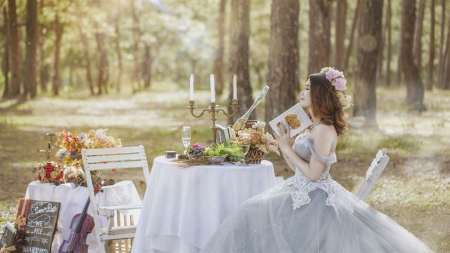 The image shows a bride wearing a light blue gown sitting at an elegantly set table in a peaceful forest. The table features flowers, candelabra, and champagne glass while soft sunlight illuminates the scene creating a serene atmosphere fitting for an intimate wedding. A nearby chair adorned with colorful flowers adds charm to this enchanting setting designed for couples dreaming of their unique celebration in Dubai.
