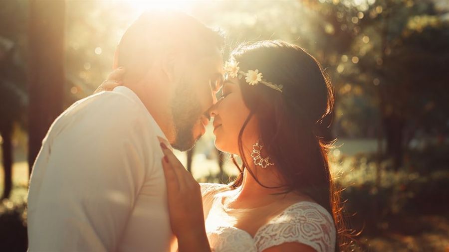 The image shows a couple warmly embracing outdoors in soft sunlight. The man wears a white shirt while the woman is dressed in a beautiful lace gown adorned with flowers. Their joyful expressions portray love and closeness, surrounded by blurred greenery that adds serenity to the scene. The lighting creates a dreamy effect hinting at a magical moment just before their kiss. This romantic setting captures the essence of weddings in Dubai and Abu Dhabi beautifully.