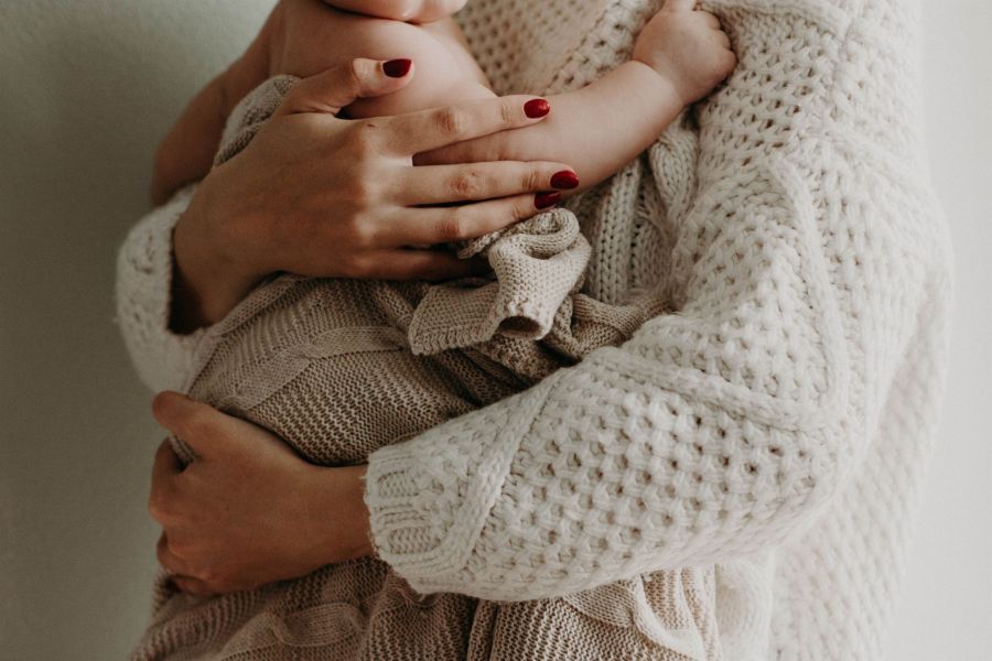 The image depicts a close-up of an adult holding a baby tightly in a cozy embrace. Both are dressed in soft knit garments in neutral colors with the adult displaying well-groomed red nails. The baby is swaddled in a knitted blanket, showcasing warmth and affection. The minimalistic background highlights this loving connection beautifully, creating a soothing atmosphere that emphasizes their bond.
